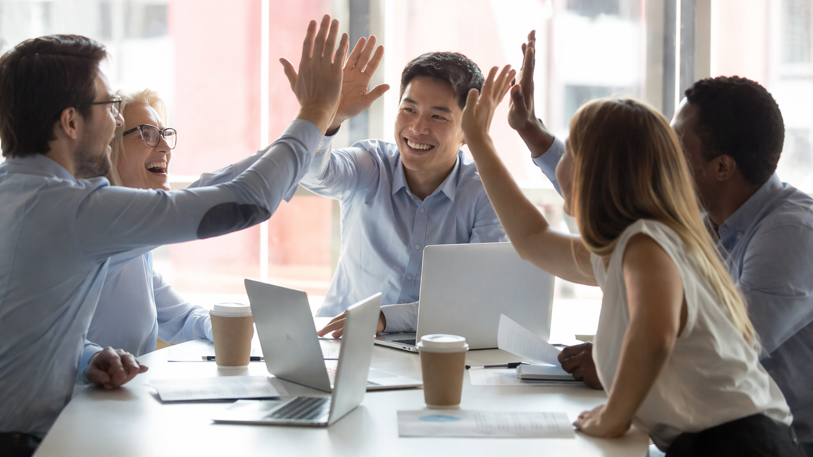 Smiling group of employees give each other high fives in well-lit conference room