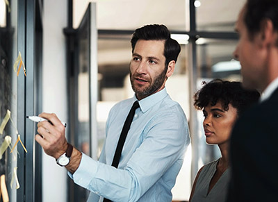 Man in shirt and tie points to a board while instructing 2 others.